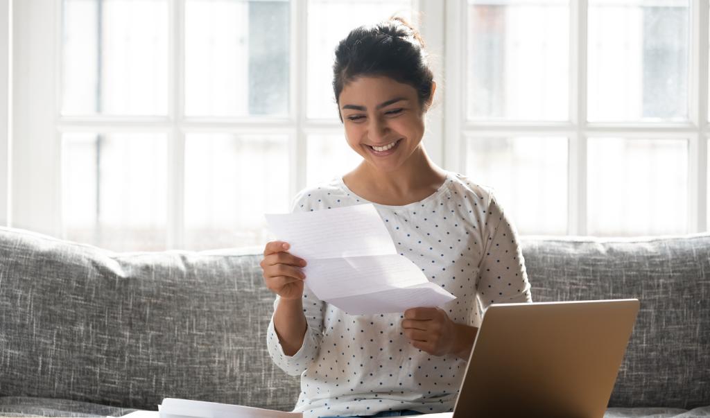 Indian ethnicity woman sitting on couch at home reading paper notice receive good news feels happy, cheerful student female looking at document enjoy exam results or college admission letter concept